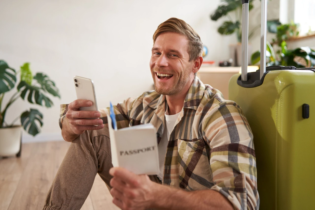 Portrait of smiling handsome tourist, man holding passport and flight tickets, sitting with suitcase. Tourism and travelling concept